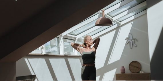 A woman standing in a sunlit attic, arms behind her head, exuding tranquility.